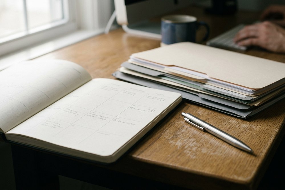 Desk planner with renewal-reminder notes in pencil and a stack of folders beside a brushed-steel pen.